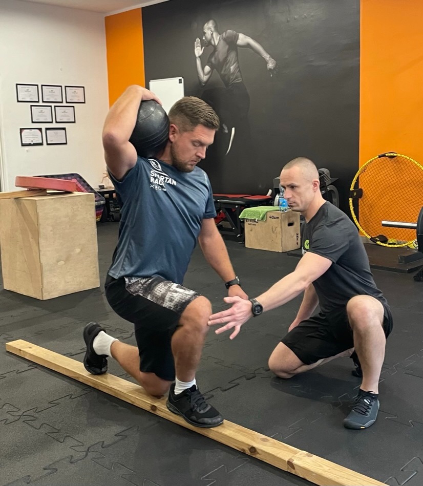 Mężczyzna wykonuje wypad z obciążeniem (piłką lekarską) na drewnianej belce pod okiem trenera w siłowni; na ścianie grafika z sylwetką biegacza.