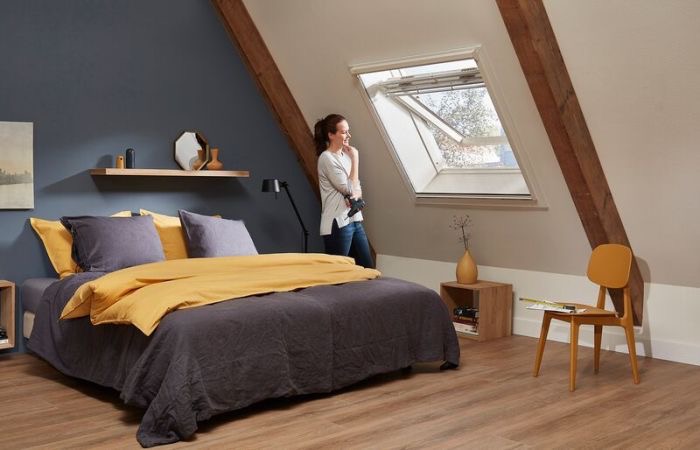 Kobieta przy oknie dachowym z moskitierą rolowaną, w sypialni na poddaszu z drewnianymi belkami i łóżkiem w kolorach szarości i żółci.