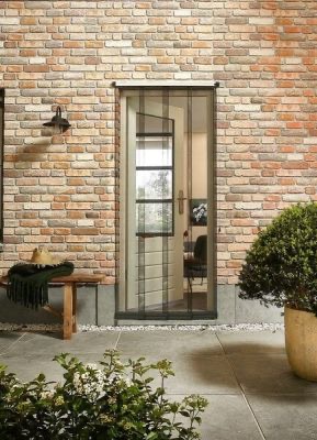 Drzwi wejściowe z siatką ochronną na magnesy, zamontowane w murze z cegły, widok z zewnątrz na taras z roślinami i ławką.