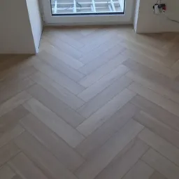 Jasne panele ułożone we wzór jodełki francuskiej, widoczne przy oknie z drabiną na zewnątrz i wystającymi kablami elektrycznymi.