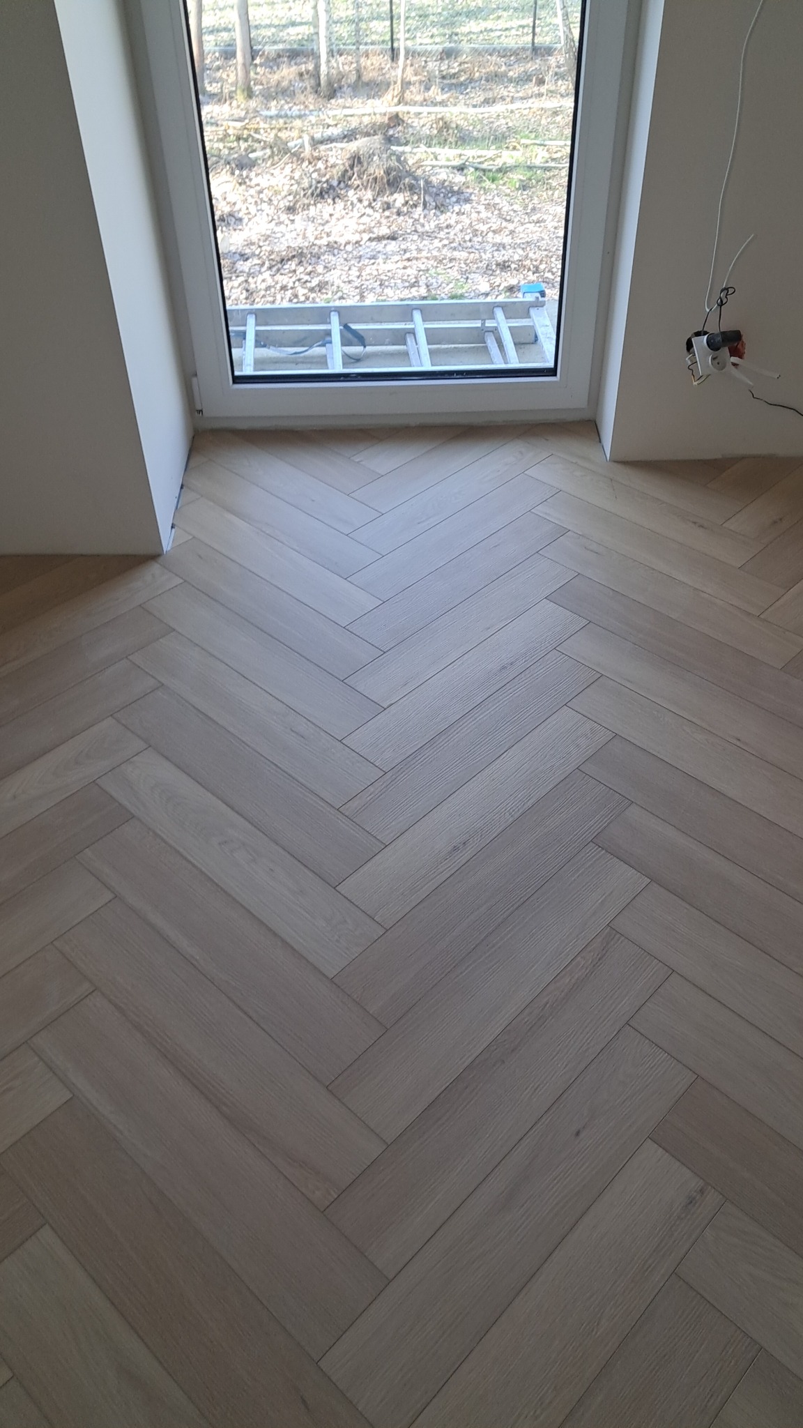 Jasne panele ułożone we wzór jodełki francuskiej, widoczne przy oknie z drabiną na zewnątrz i wystającymi kablami elektrycznymi.