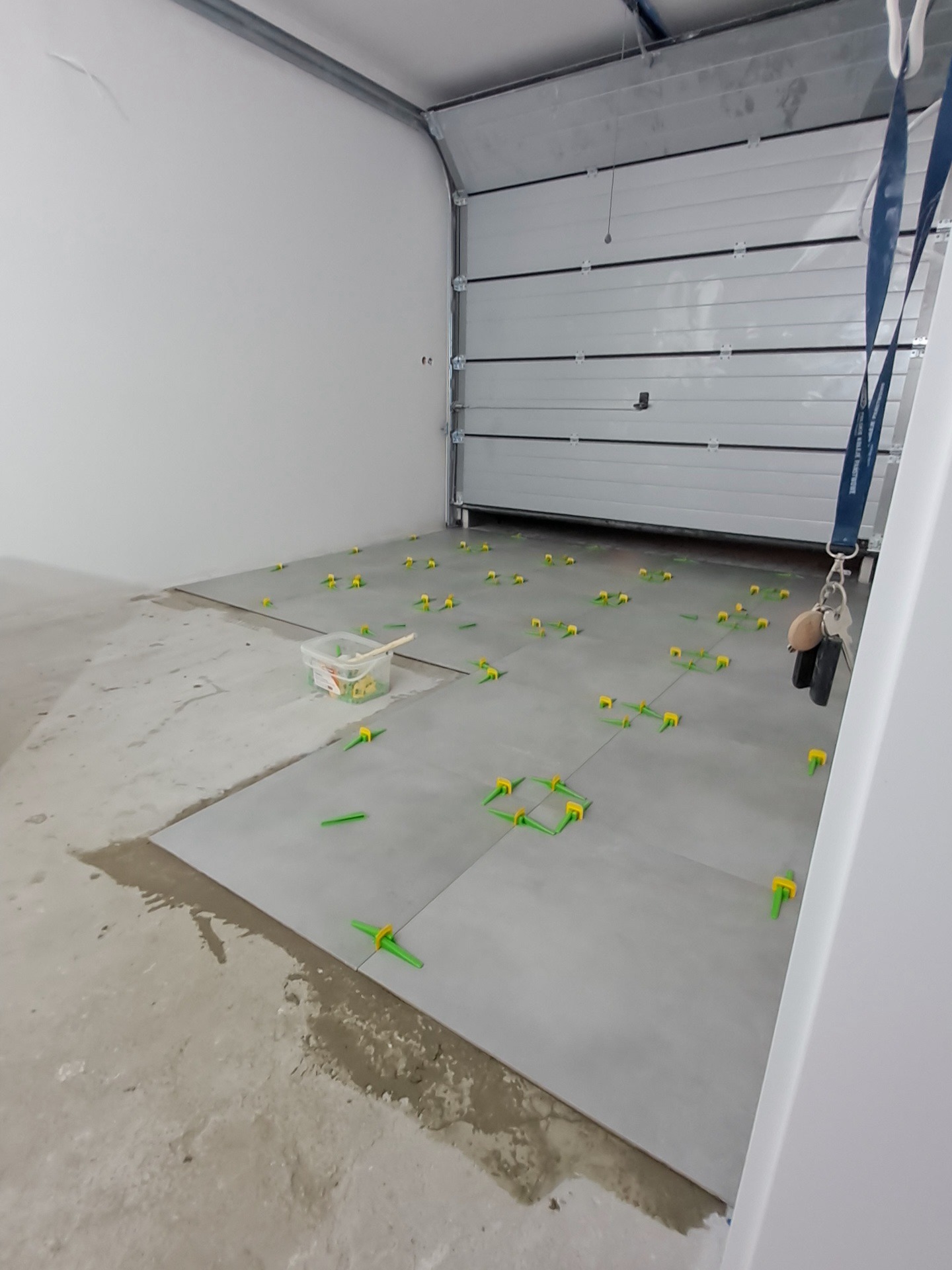 Szare płytki podłogowe w garażu, w trakcie układania, z użyciem zielonych klinów poziomujących; widoczne ślady zaprawy i otwarte drzwi garażowe.