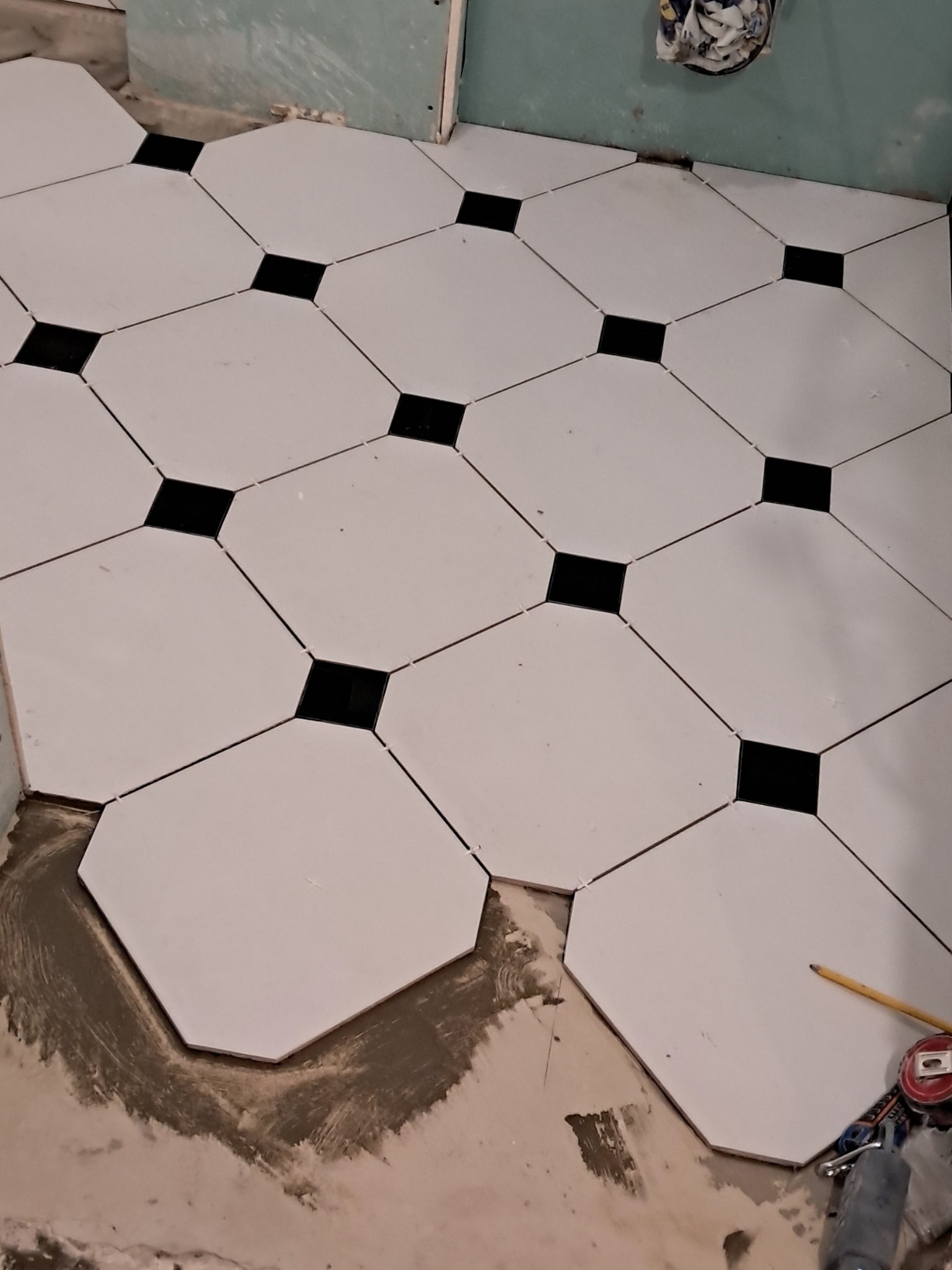 Układanie podłogi z białych, ośmiokątnych płytek z czarnymi kwadratami w narożach; widoczne narzędzia glazurnicze i zaprawa.