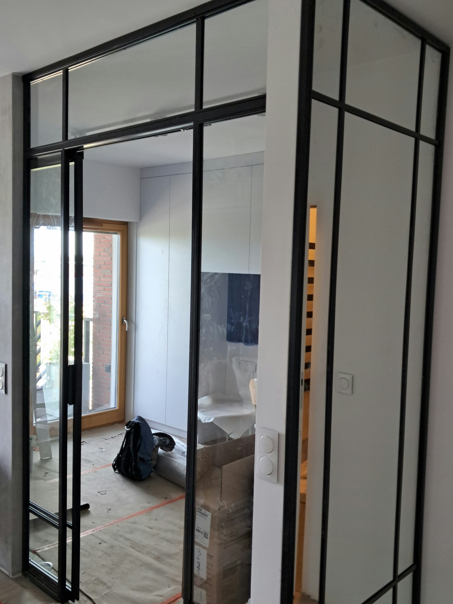 Szklana ścianka działowa z czarnymi profilami, przesuwne drzwi, widoczne zabezpieczenia podłogi podczas prac wykończeniowych.