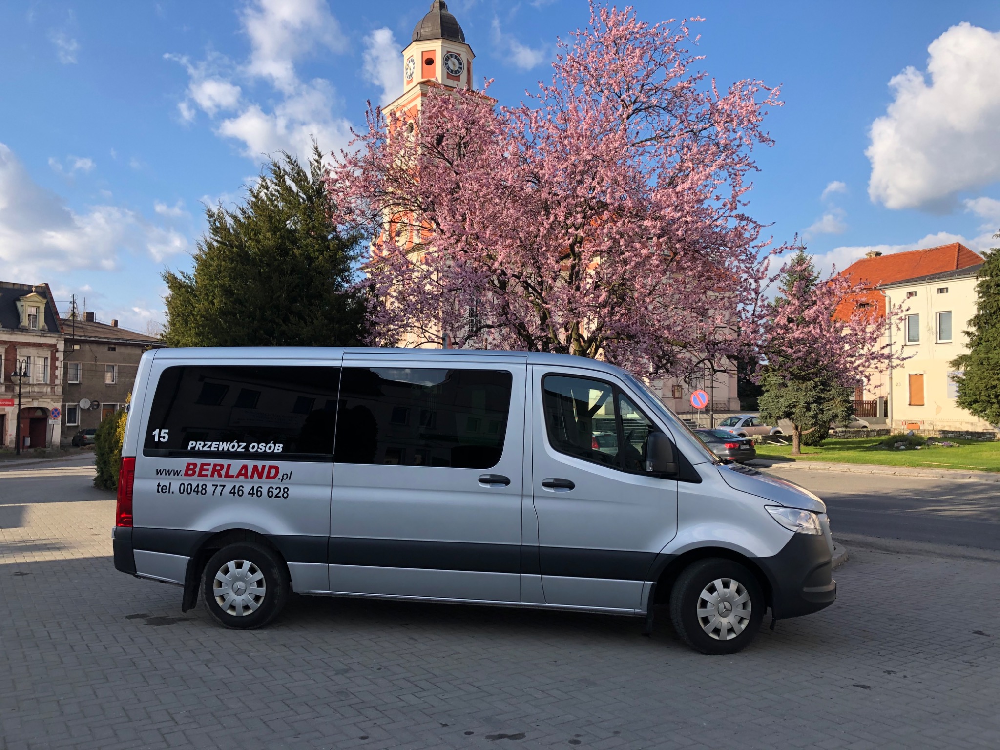 Srebrny bus z napisem 'Przewóz Osób' na tle kwitnącego drzewa i wieży zegarowej.