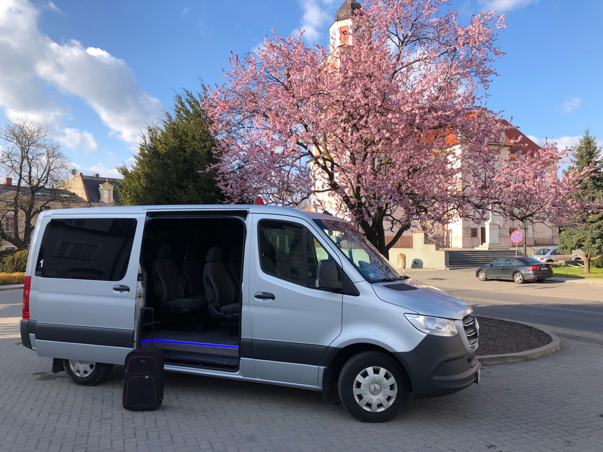 Srebrny bus z otwartymi drzwiami na tle kwitnącego drzewa i budynku z wieżą zegarową; obok stoi czarna walizka na kółkach.