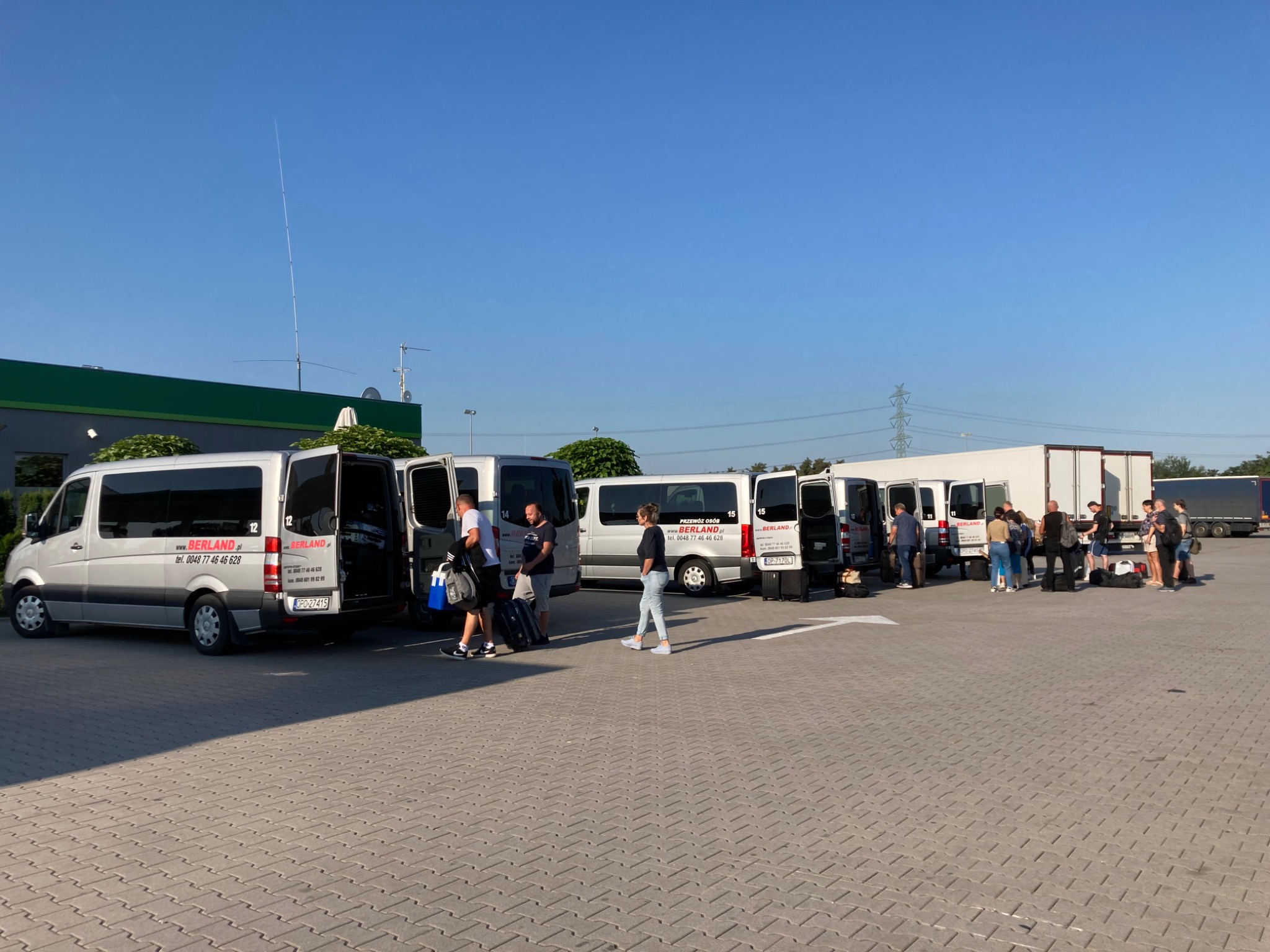 Srebrne busy firmy BERLAND z otwartymi drzwiami, pasażerowie z bagażami na parkingu przed budynkiem w słoneczny dzień.
