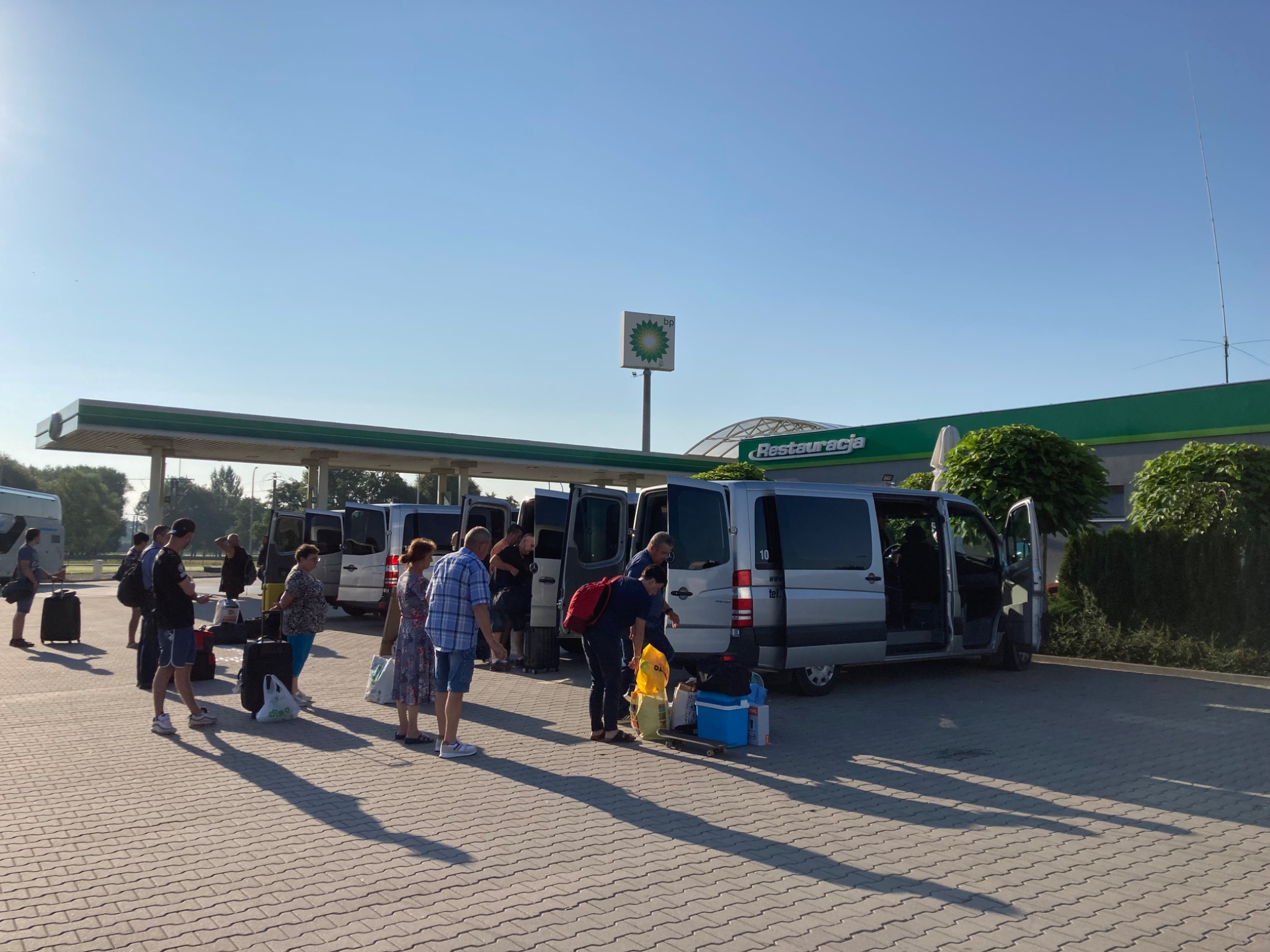 Grupa pasażerów przy srebrnych busach na parkingu stacji benzynowej BP, pakujących bagaże do otwartych bagażników w słoneczny dzień.