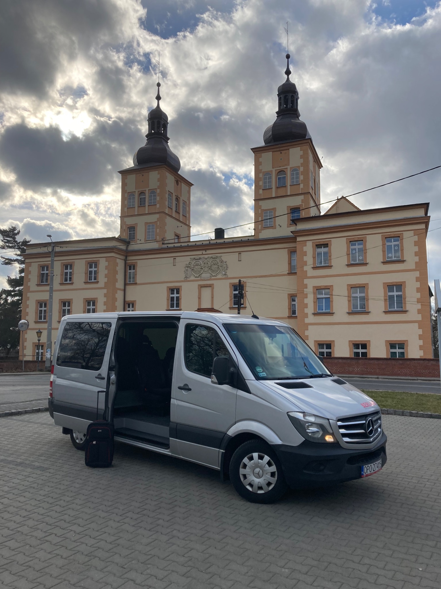 Srebrny bus z otwartymi drzwiami na tle budynku z dwiema wieżami, obok busa stoi czarna torba podróżna.