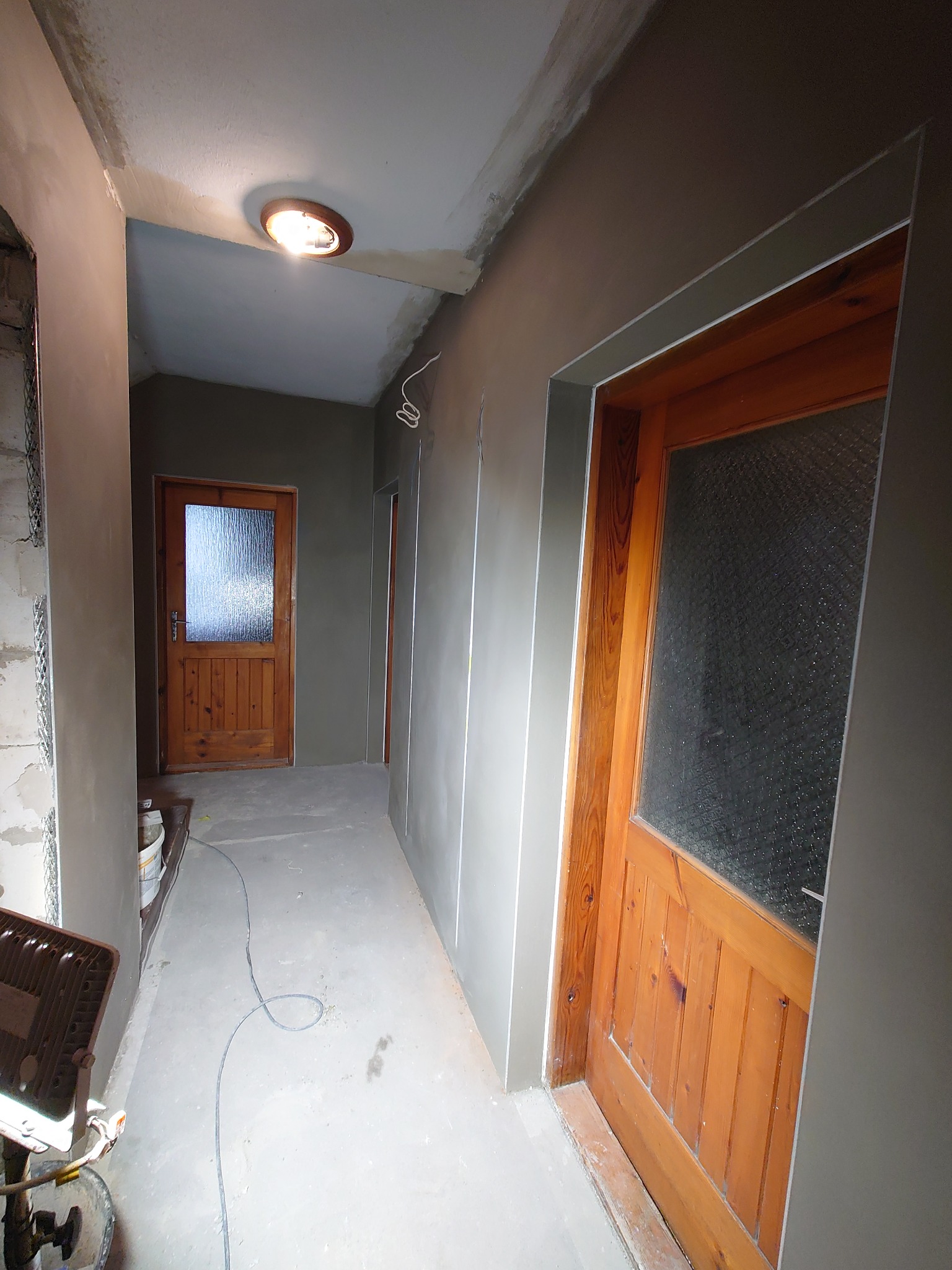 Korytarz w trakcie prac wykończeniowych: ściany pokryte świeżym tynkiem, widoczne drewniane drzwi z szybą, kabel zwisający ze ściany. Oświetlenie sufitowe.