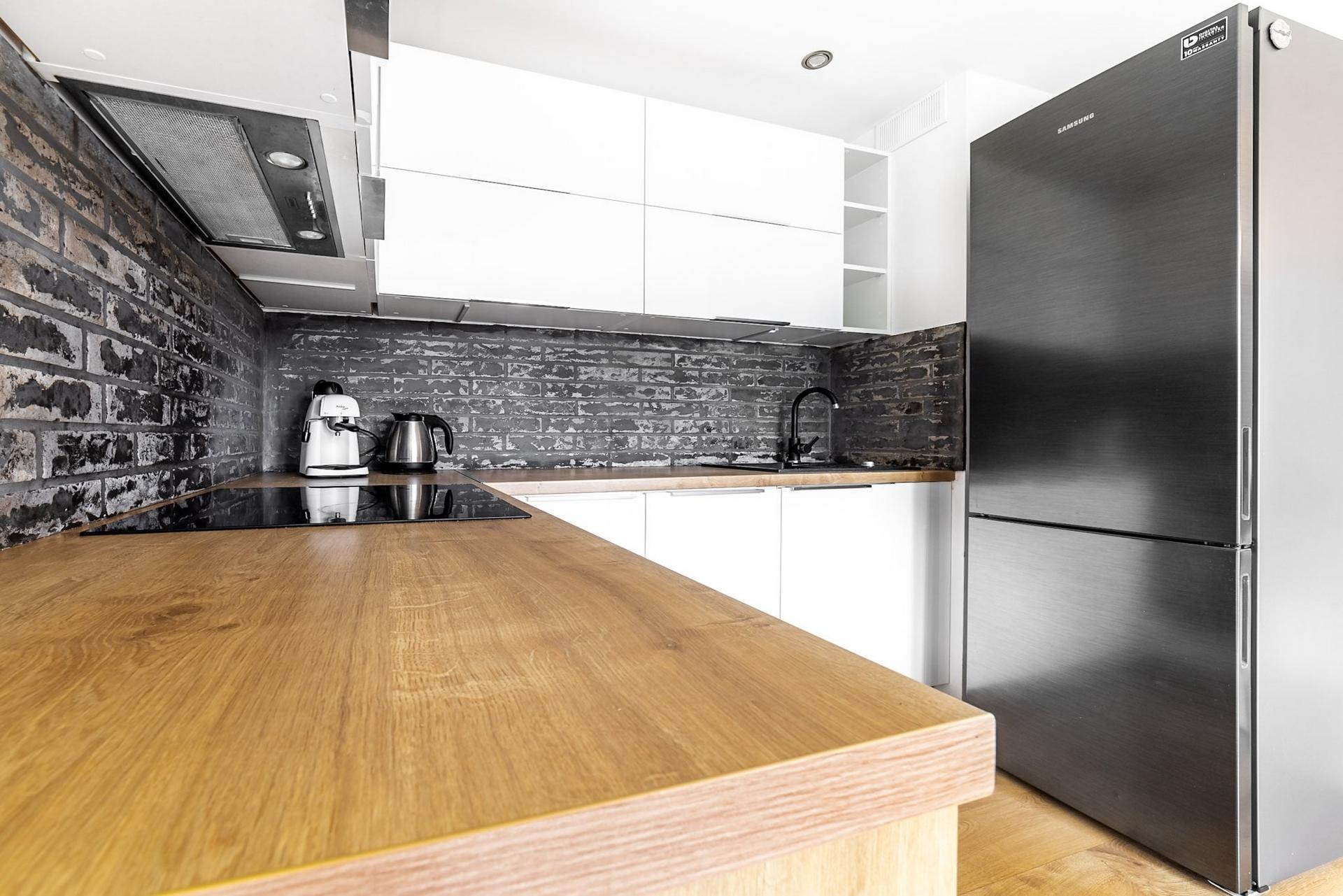 Nowoczesna kuchnia w Krakowie z blatem z jasnego drewna, ceglaną ścianą w odcieniach szarości i czarną armaturą, uzupełniona o białe szafki i srebrną lodówkę Samsung.