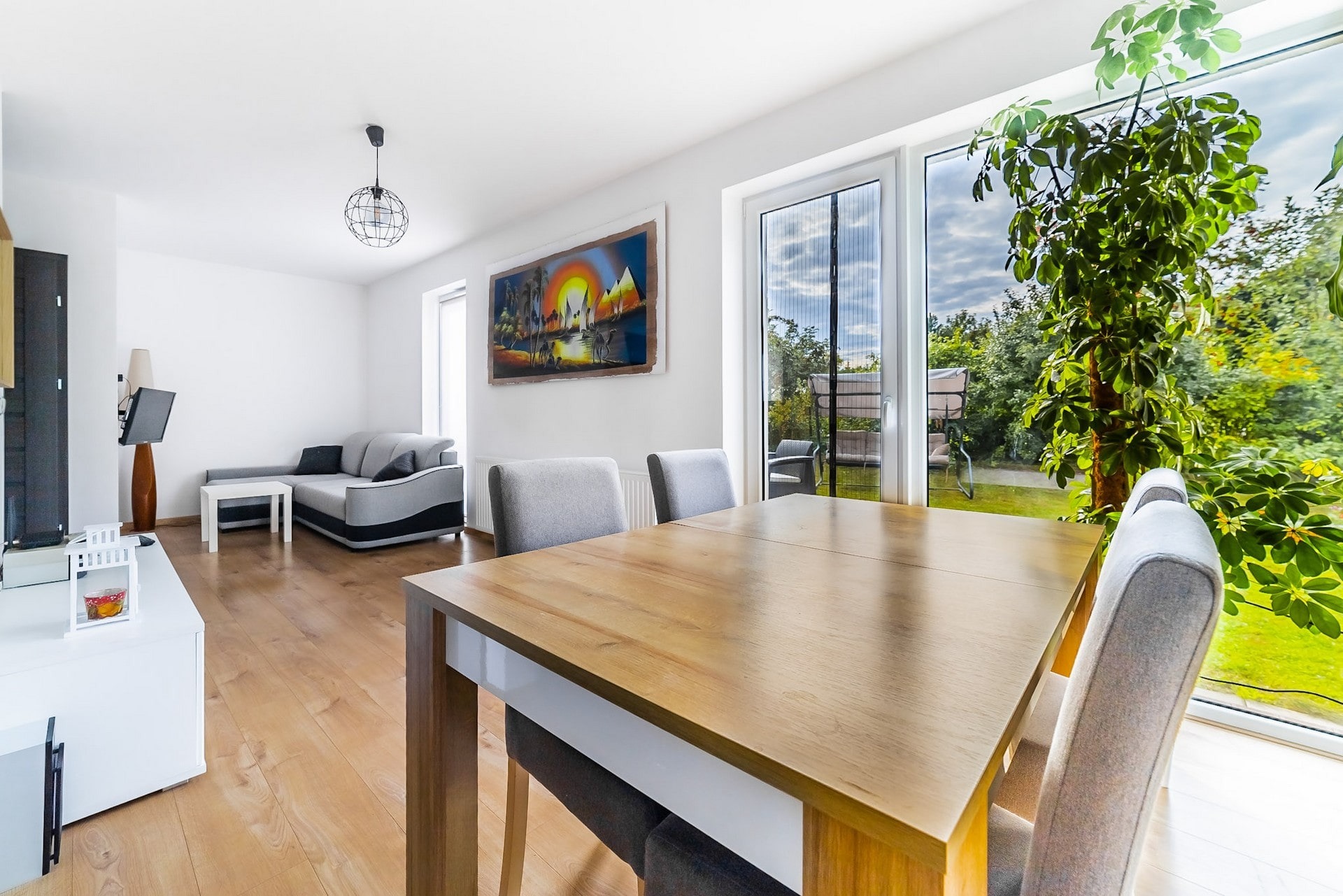 Jasne wnętrze salonu z drewnianym stołem jadalnym na pierwszym planie, szara sofa i telewizor w tle, widok na ogród przez duże okno z moskitierą.