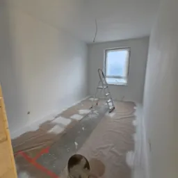 Pok&oacute;j w trakcie malowania na biało, podłoga zabezpieczona folią z czerwoną taśmą, drabina aluminiowa i wiadro z farbą widoczne na pierwszym planie.