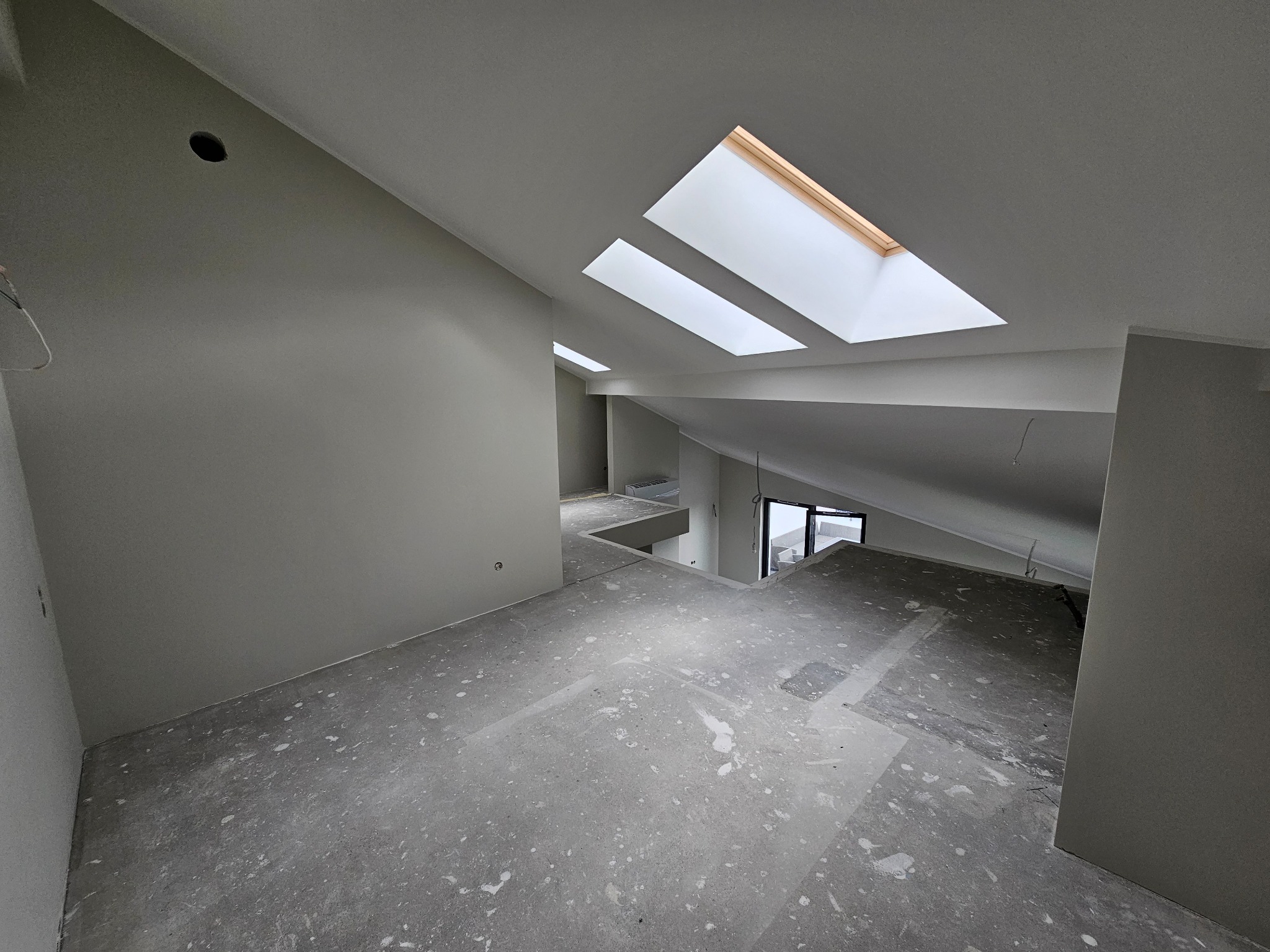 Wnętrze domu w trakcie prac wykończeniowych, widoczne surowe ściany w jasnym odcieniu szarości, trzy okna dachowe wpuszczające naturalne światło i podłoga z widocznymi śladami po pracach budowlanych.