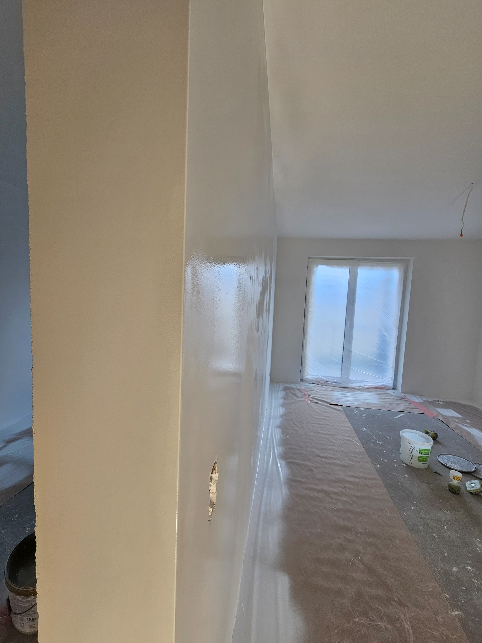 Świeżo wyszpachlowana ściana z widocznym ubytkiem do naprawy, zabezpieczona folią okno i podłoga, widoczne narzędzia malarskie i wiadro z farbą.