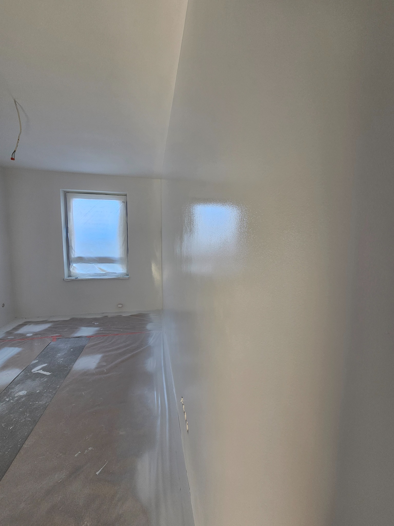 Wnętrze pokoju po świeżym pomalowaniu na biało, okno zabezpieczone folią, podłoga zakryta folią ochronną z widocznymi fragmentami szarych płyt.