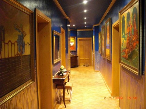 Długi korytarz w rezydencji, ściany w odcieniach niebieskiego i żółtego, zdobione obrazami w złotych ramach, widoczny stolik z kwiatami i mały taboret.