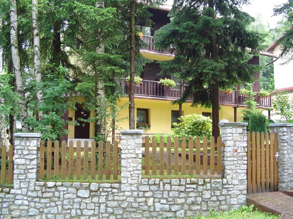 Pensjonat w Karpaczu: dwupiętrowy budynek z żółtą elewacją, drewnianymi balkonami z kwiatami, otoczony drzewami i kamienno-drewnianym ogrodzeniem.