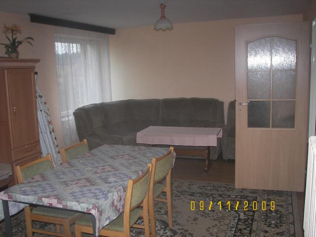 Jasne wnętrze pokoju z dużym stołem przykrytym wzorzystym obrusem, krzesłami z zielonymi obiciami, zieloną sofą i drewnianą szafą. Na podłodze dywan z datą wykonania zdjęcia.