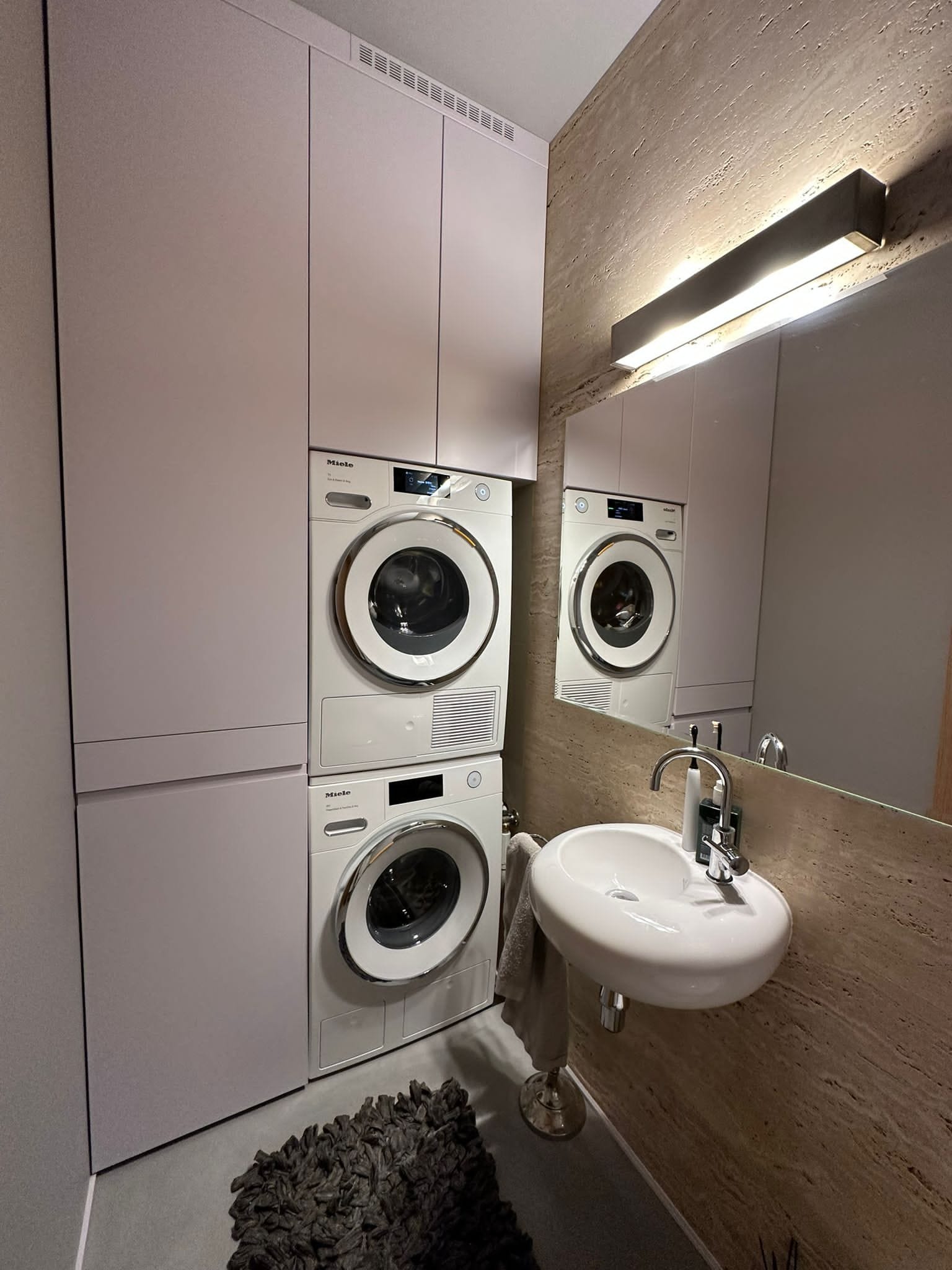 Nowoczesna łazienka z pralką i suszarką Miele w zabudowie meblowej w kolorze pudrowego różu, umywalka nablatowa na tle ściany z trawertynu i lustra z oświetleniem.