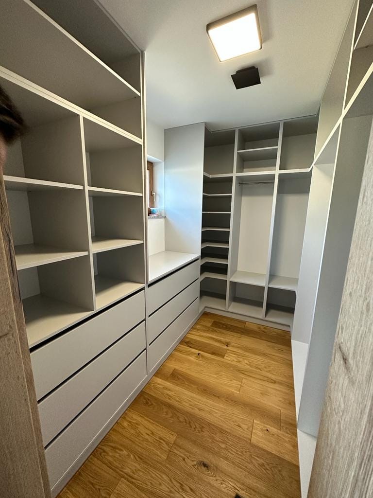 Jasnoszara garderoba wnękowa z drewnianą podłogą, oświetleniem sufitowym i półkami oraz szufladami.