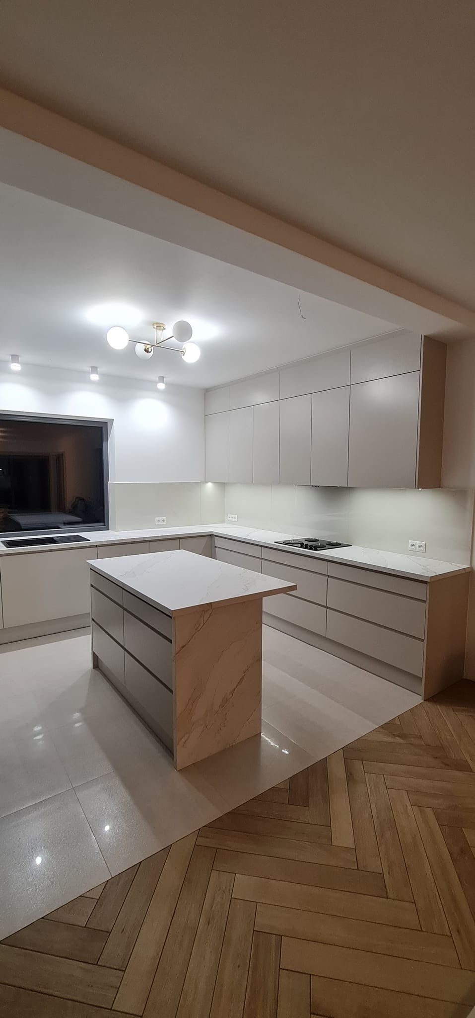 Jasna kuchnia z wyspą, blaty imitujące marmur, nowoczesne oświetlenie punktowe i lampa wisząca, połączenie płytek podłogowych i parkietu w jodełkę.