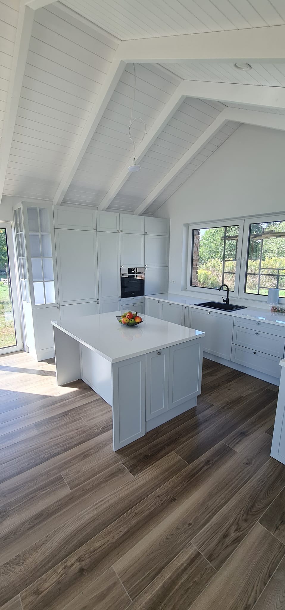 Jasna kuchnia z wyspą i drewnopodobną podłogą; na wyspie misa z owocami, widok na zewnątrz przez okno.