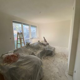 Patryk kindel - Pokój w trakcie malowania: zabezpieczony mebel folią, drabina przy oknie, ślady farby na podłodze. Widoczna szlifierka do gładzi i pomalowane ściany.