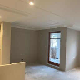 Patryk kindel - Pokój po malowaniu ścian na beżowo, z dekoracyjnymi listwami sufitowymi i gniazdkami elektrycznymi w ramce. Widoczny odkurzacz przemysłowy i surowa podłoga.