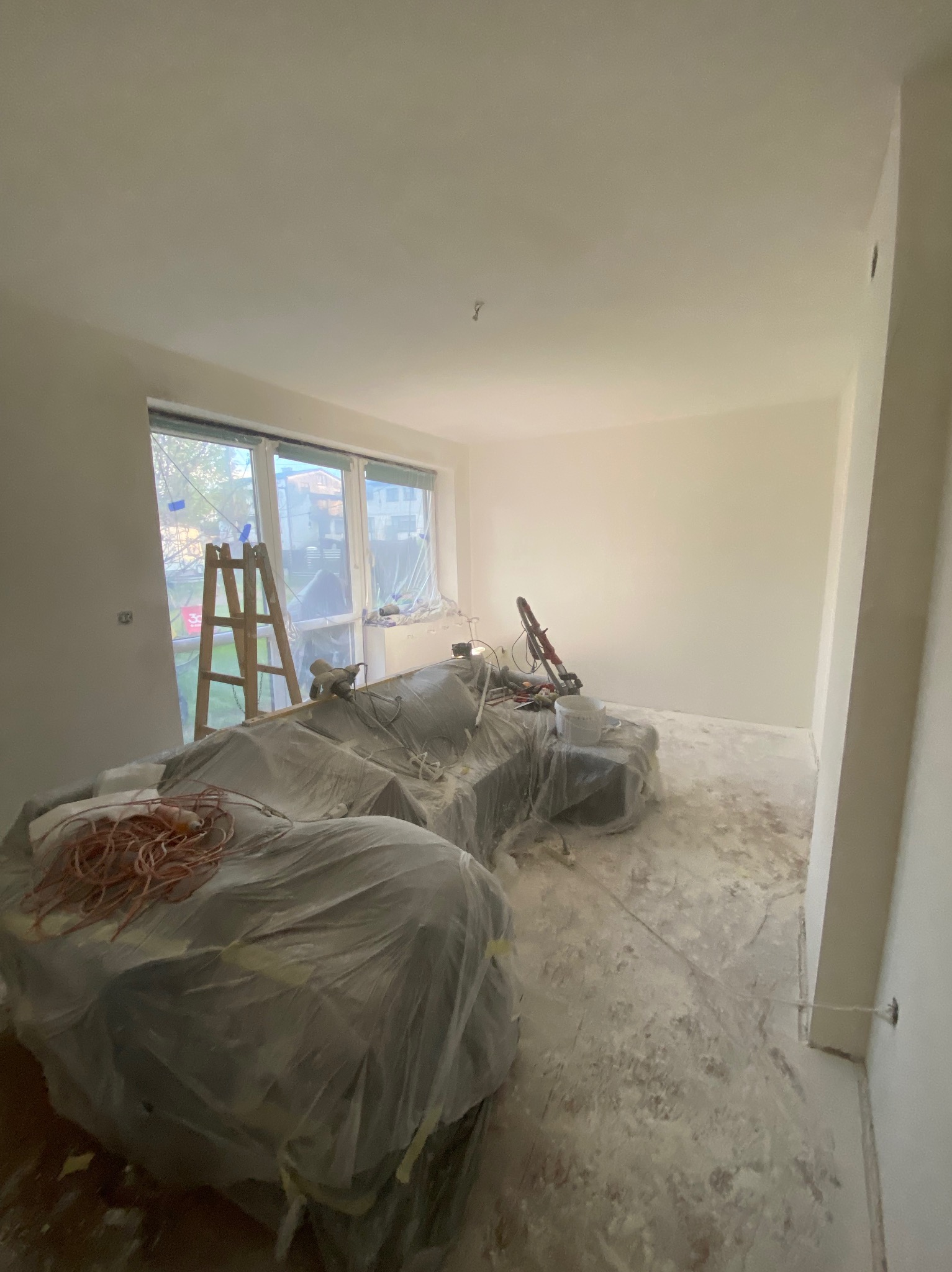 Pokój w trakcie malowania: zabezpieczony mebel folią, drabina przy oknie, ślady farby na podłodze. Widoczna szlifierka do gładzi i pomalowane ściany.