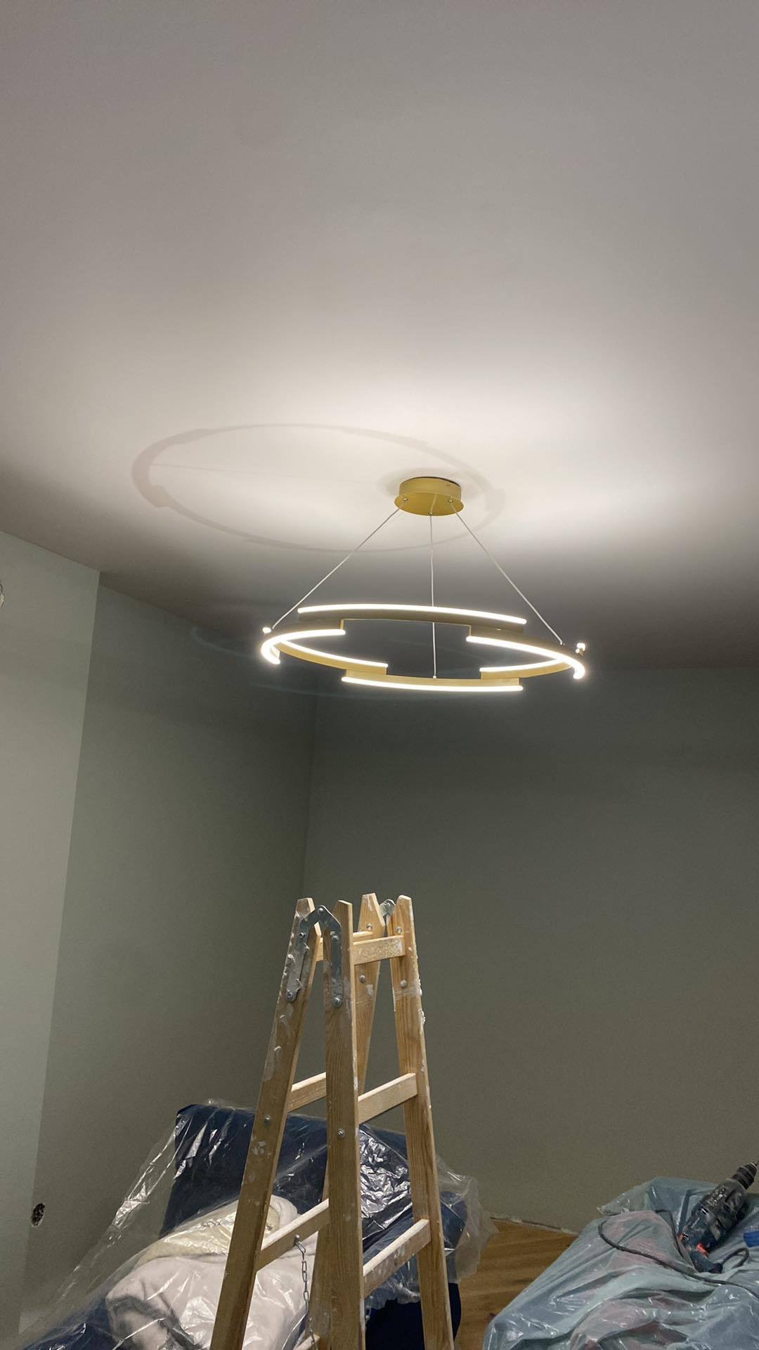Nowoczesna lampa wisząca w kształcie pierścienia, zamontowana na świeżo pomalowanym suficie. W tle ściana w odcieniach szarości i drabina malarska.