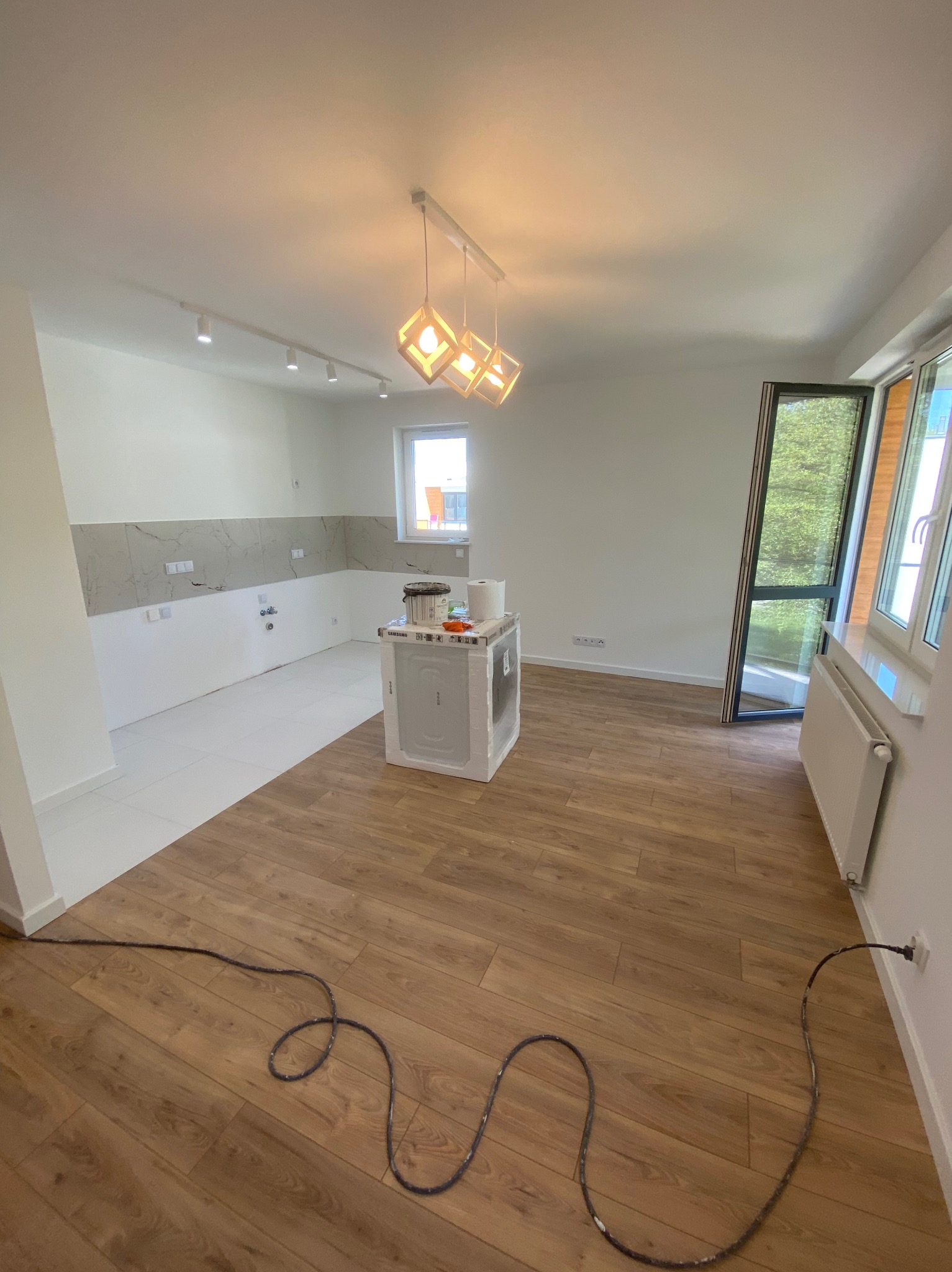 Jasne wnętrze po remoncie: drewniana podłoga, białe ściany z szarym pasem płytek w kuchni, nowoczesne oświetlenie i okno z widokiem na zieleń. Widoczne akcesoria malarskie.