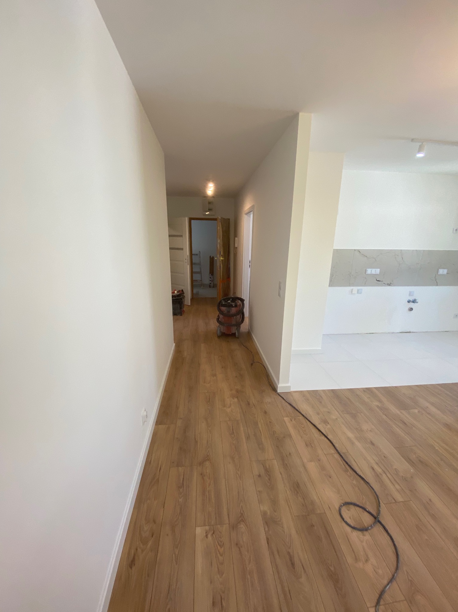 Jasne, świeżo pomalowane ściany w korytarzu i kuchni. Na podłodze panele. Widoczny odkurzacz przemysłowy i kabel. Kuchnia z białymi płytkami i szarym pasem nad blatem.