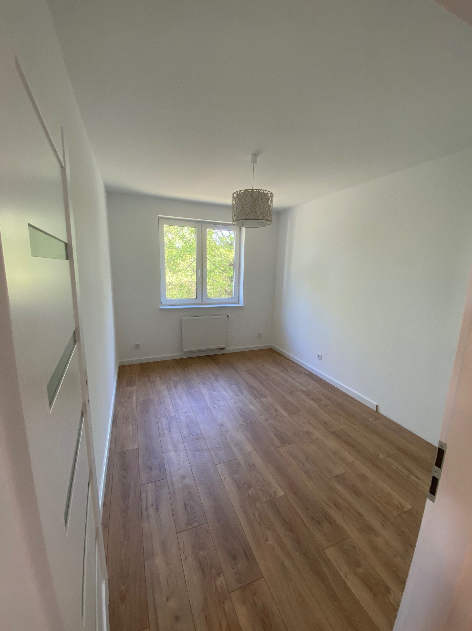 Jasny pokój po remoncie: drewniane panele podłogowe, białe ściany, okno z widokiem na zieleń. Minimalistyczny wystrój z lampą sufitową. Widok na pokój zza otwartych drzwi.