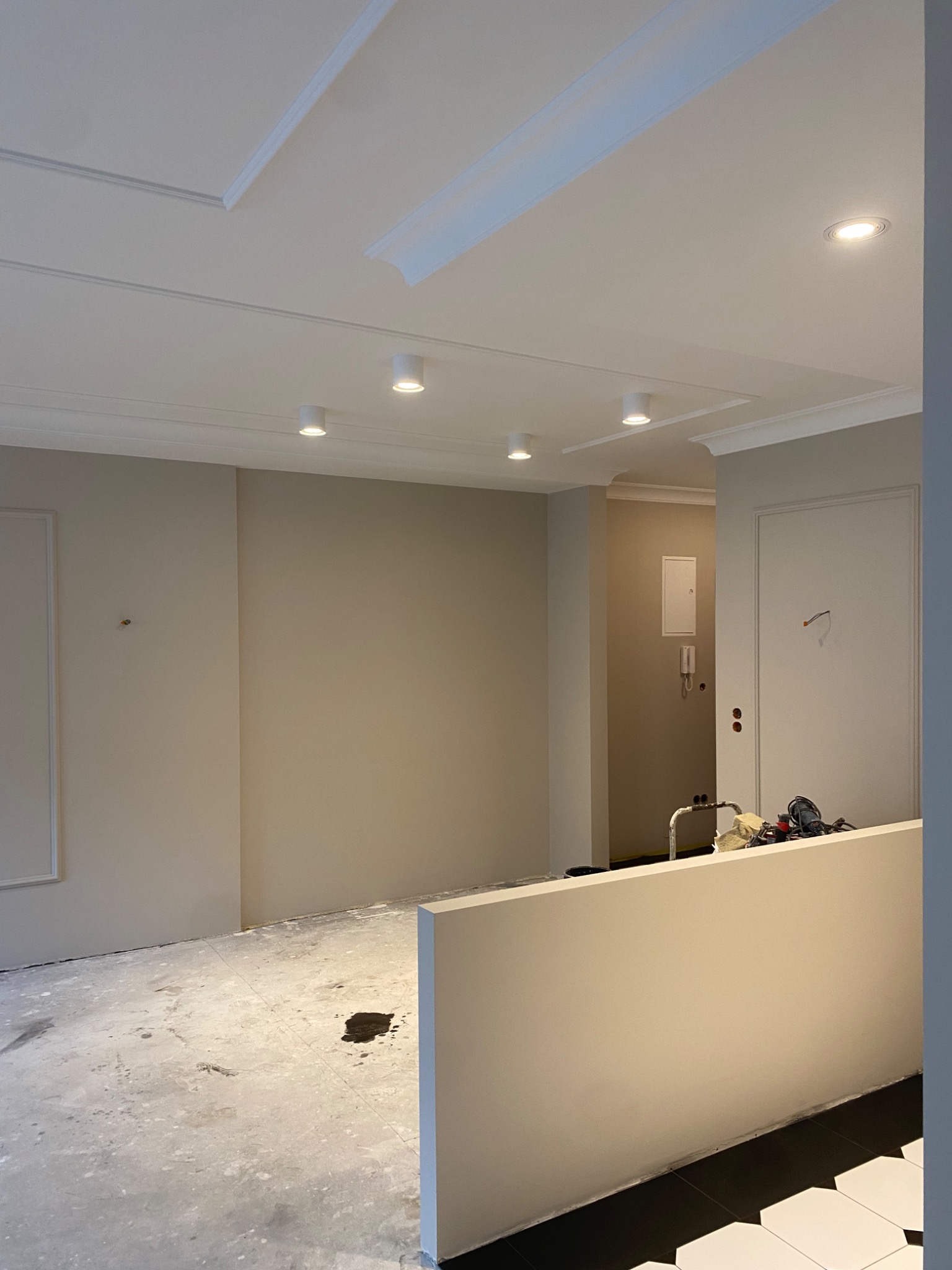 Jasne wnętrze w trakcie wykańczania: pomalowane ściany w beżowym odcieniu, ozdobny sufit z oświetleniem punktowym, fragment podłogi z czarno-białą mozaiką.