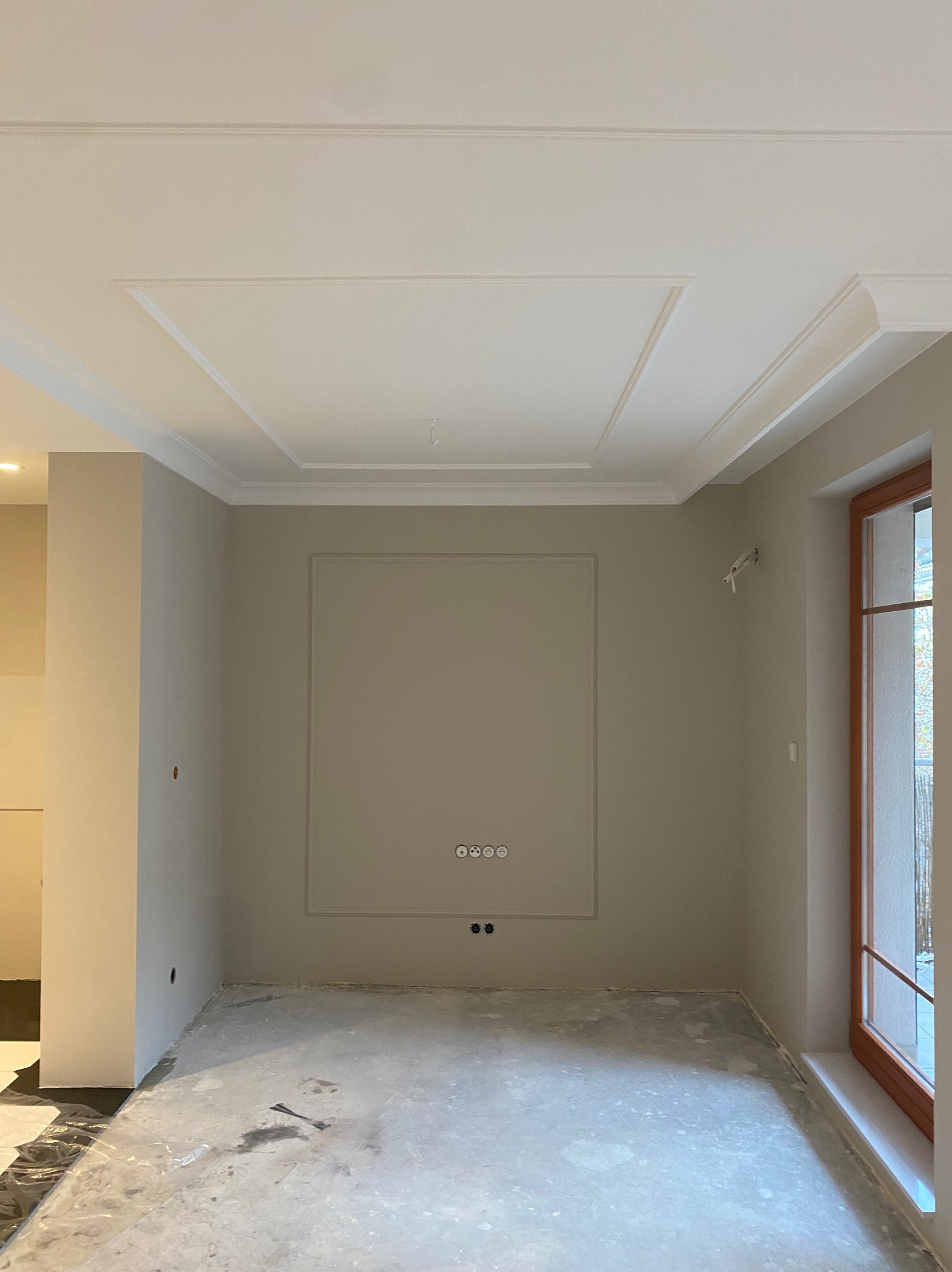 Pokój po szpachlowaniu i malowaniu na szaro, z dekoracyjną sztukaterią na ścianach i suficie. Widoczne gniazdka elektryczne i surowa podłoga przed wykończeniem.