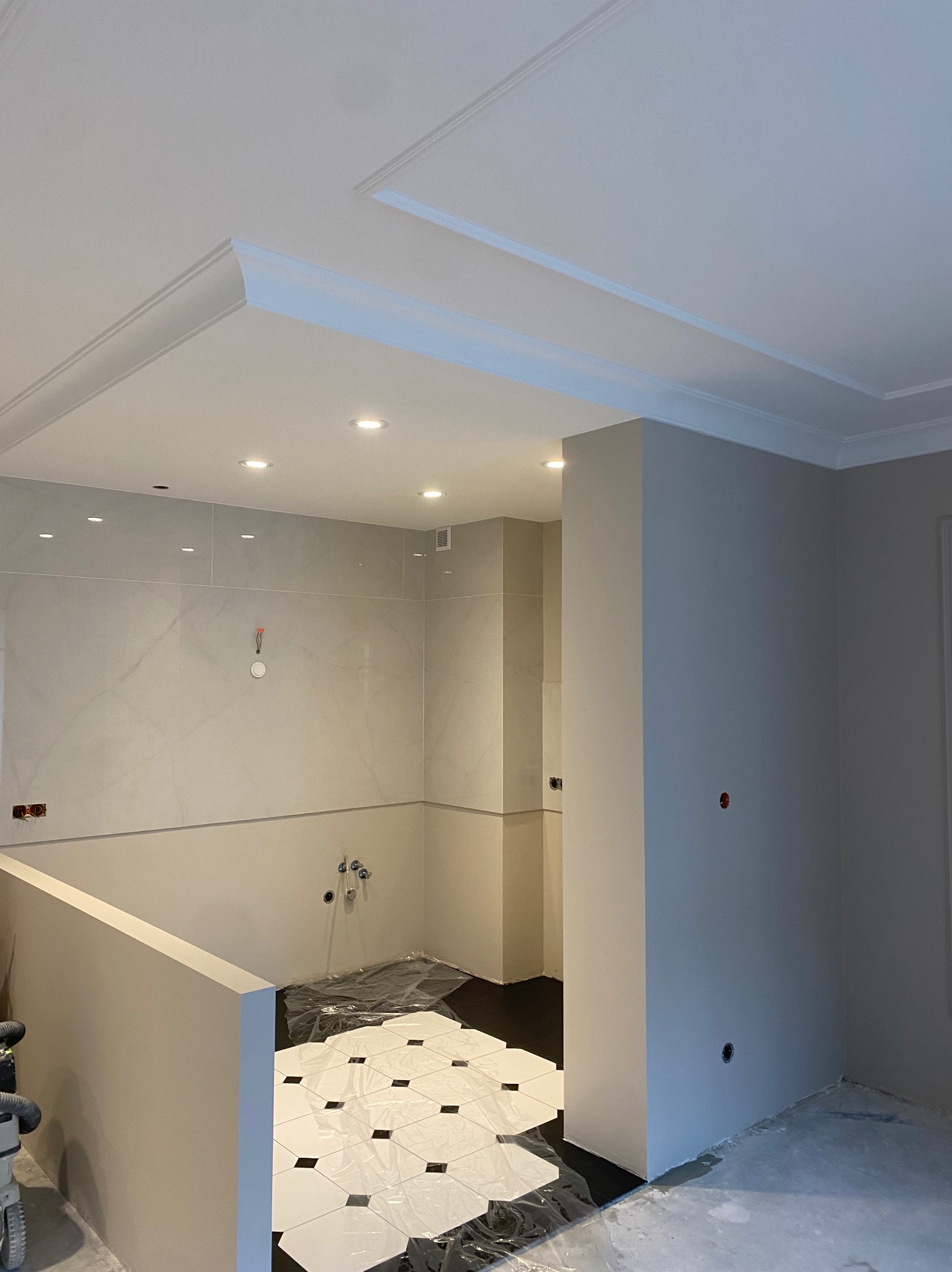 Wykończone wnętrze łazienki z marmurowymi płytkami, dekoracyjnym sufitem i czarno-białą podłogą zabezpieczoną folią. Widoczne punkty świetlne i przygotowane instalacje.