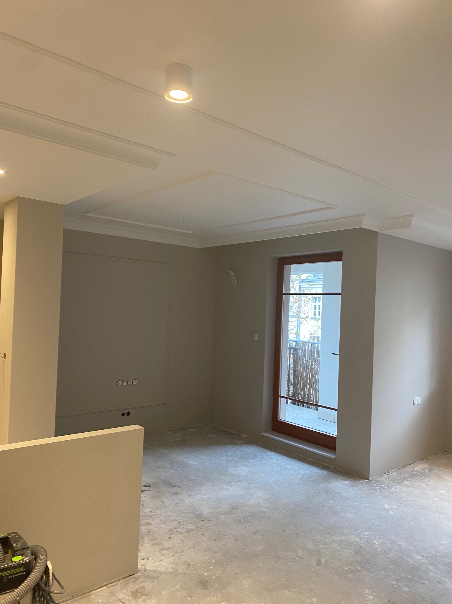 Pokój po malowaniu ścian na beżowo, z dekoracyjnymi listwami sufitowymi i gniazdkami elektrycznymi w ramce. Widoczny odkurzacz przemysłowy i surowa podłoga.
