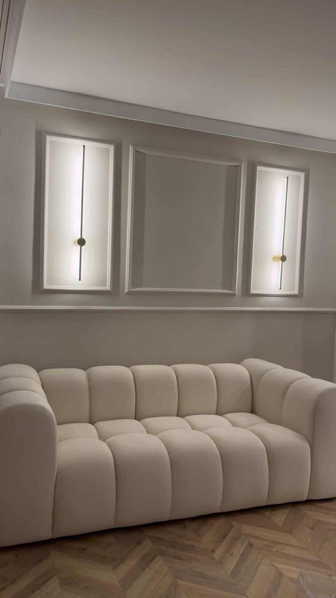 Eleganckie wykończenie wnętrza: kremowa sofa na parkiecie w jodełkę, ściana z dekoracyjnymi listwami i symetrycznie rozmieszczone, podłużne lampy ścienne.