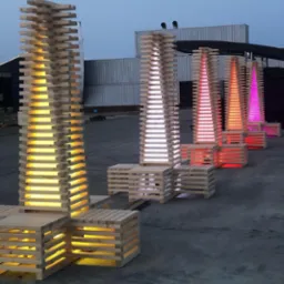 Instalacja artystyczna na zewnątrz: rzędy drewnianych wież z podświetleniem LED w kolorach żółtym, białym, czerwonym i różowym, umieszczone na betonowej powierzchni z kontenerami w tle.