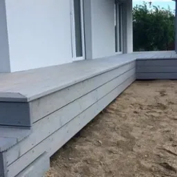 Szary taras z kompozytu drewna, prowadzący do drzwi balkonowych, z widocznymi śladami po budowie na piasku.