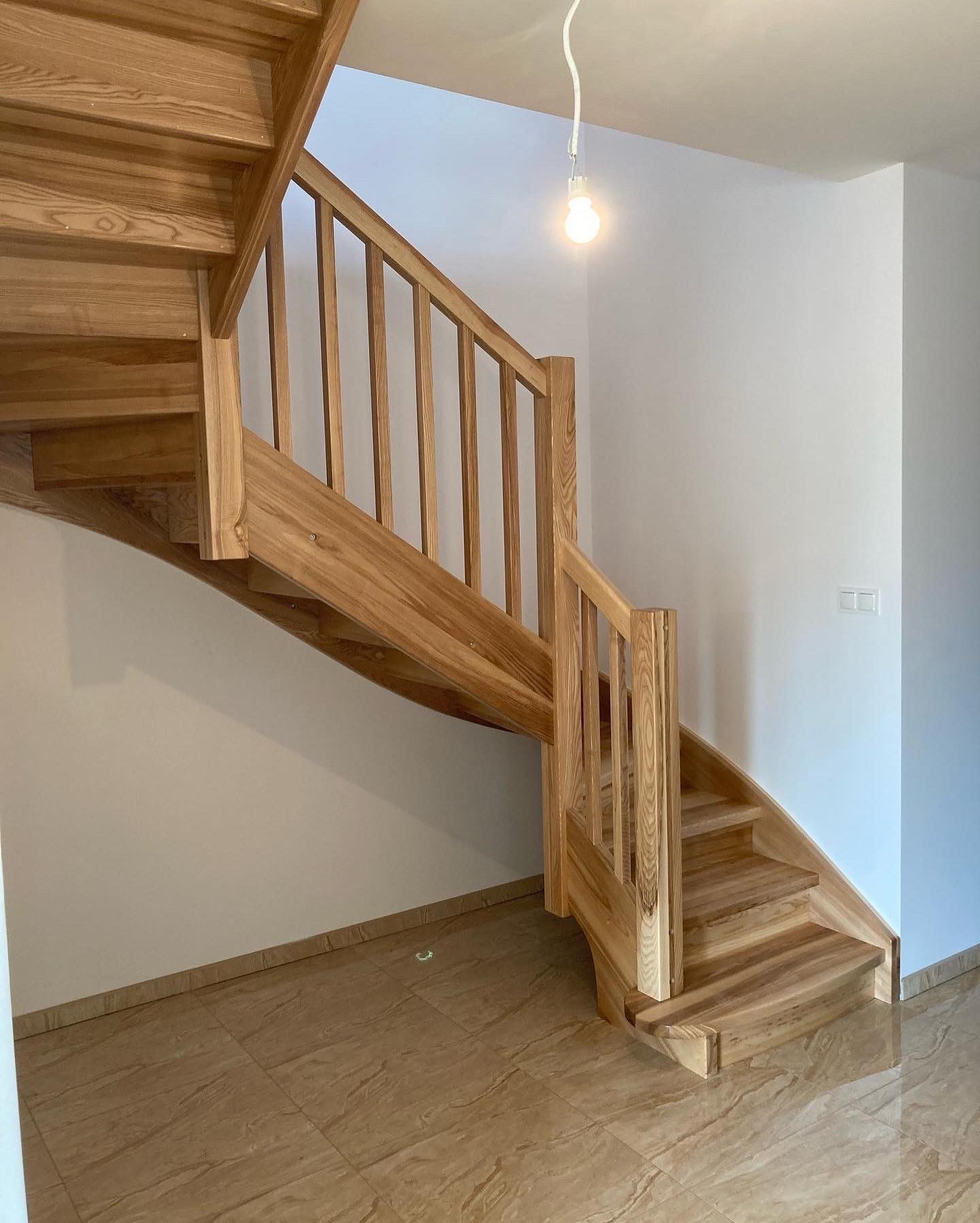 Jasne, drewniane schody z balustradą w nowoczesnym wnętrzu, widok z dołu, z widoczną żarówką na kablu zwisającą z sufitu.
