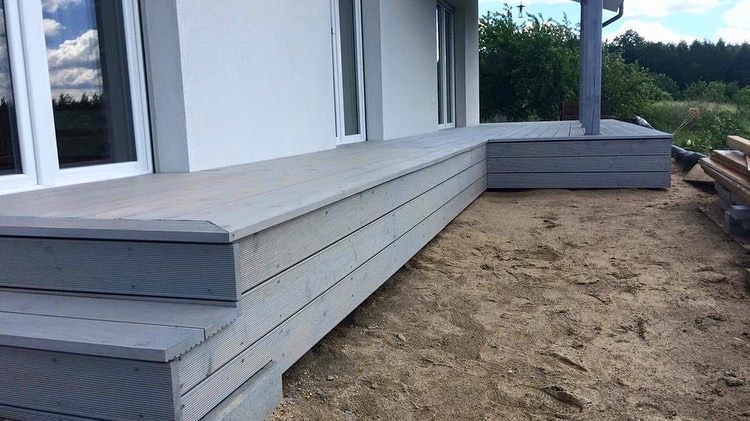 Szary taras z kompozytu drewna, prowadzący do drzwi balkonowych, z widocznymi śladami po budowie na piasku.
