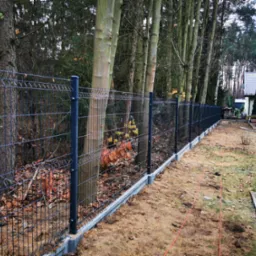 Nowo postawione, ciemne ogrodzenie panelowe na betonowej podmurówce, widoczne pomiędzy drzewami na działce, z elementami montażowymi na słupkach i pomarańczowym kablem na ziemi.
