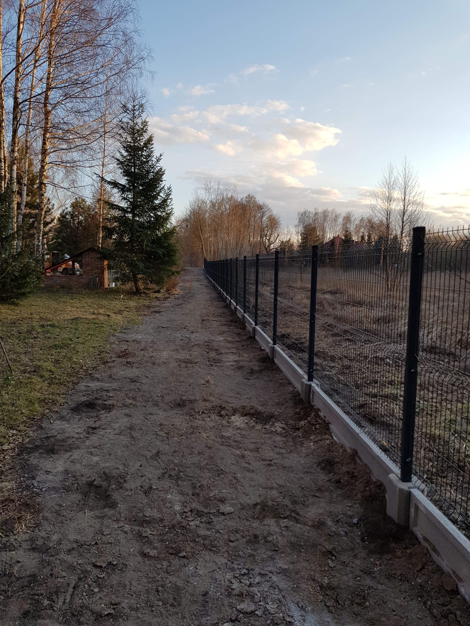 Długi fragment ogrodzenia panelowego z podmurówką betonową, widok z perspektywy ścieżki gruntowej, w tle drzewa i budynek gospodarczy, pochmurne niebo.