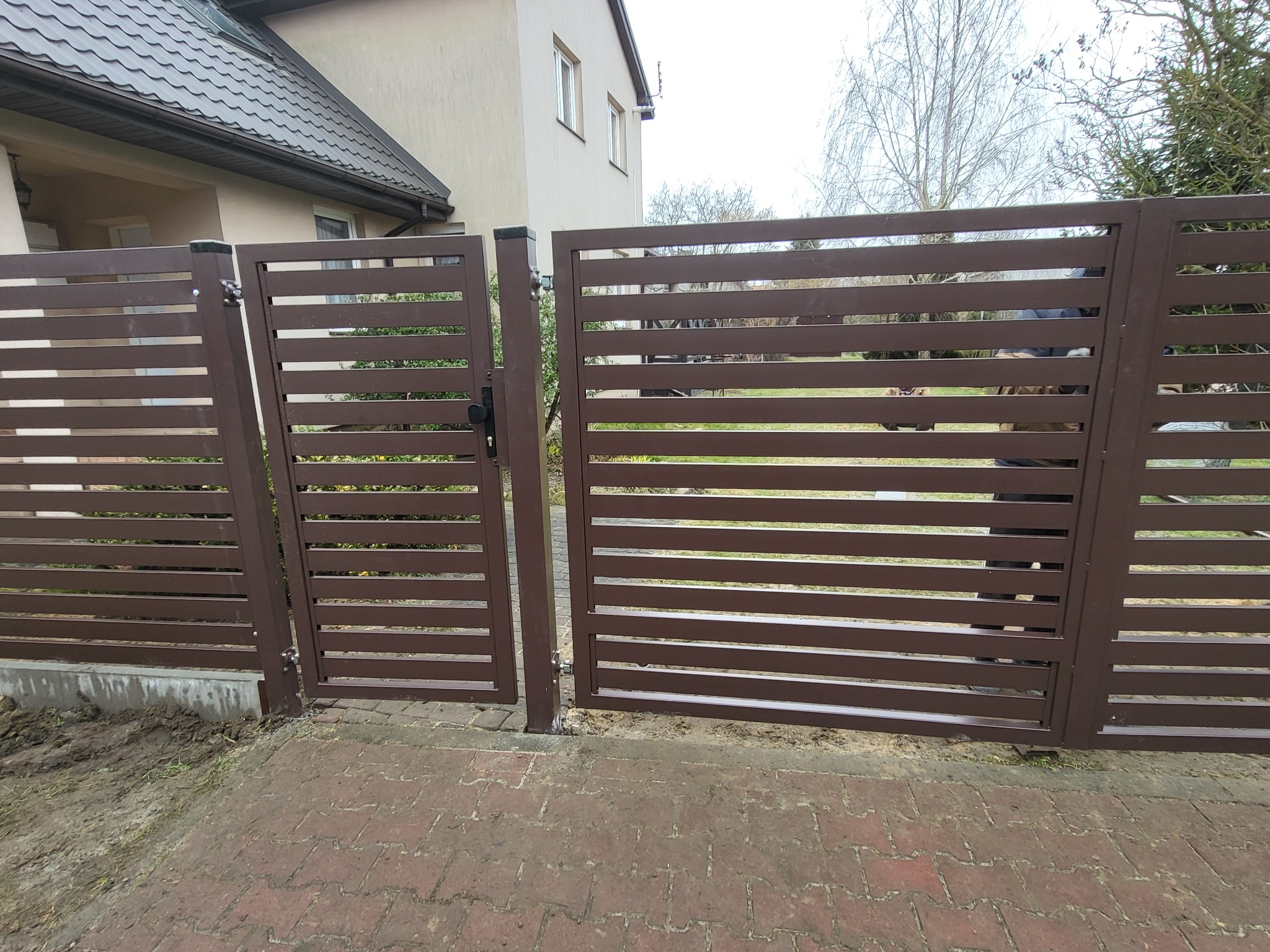 Nowoczesna brama wjazdowa z poziomych, brązowych profili metalowych, częściowo otwarta, widok na dom i drzewa w tle.