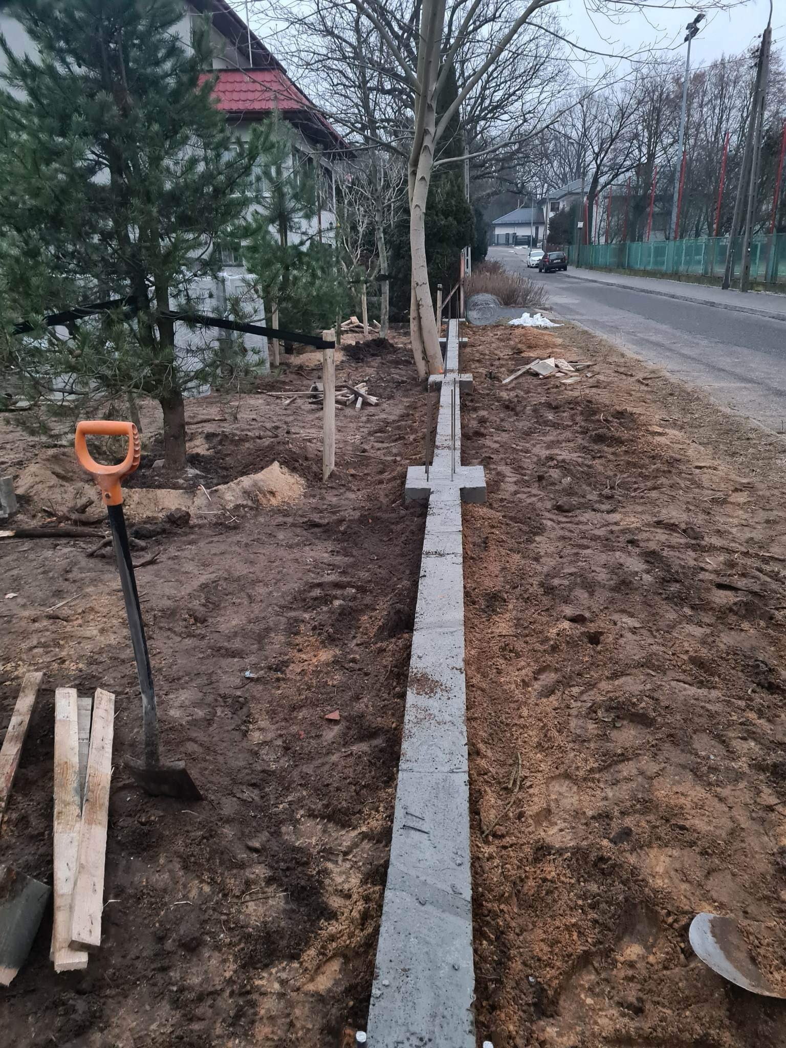 Wykopany rów pod fundament ogrodzenia betonowego z widoczną zbrojoną podstawą, szpadel wbity w ziemię, w tle dom i drzewa.
