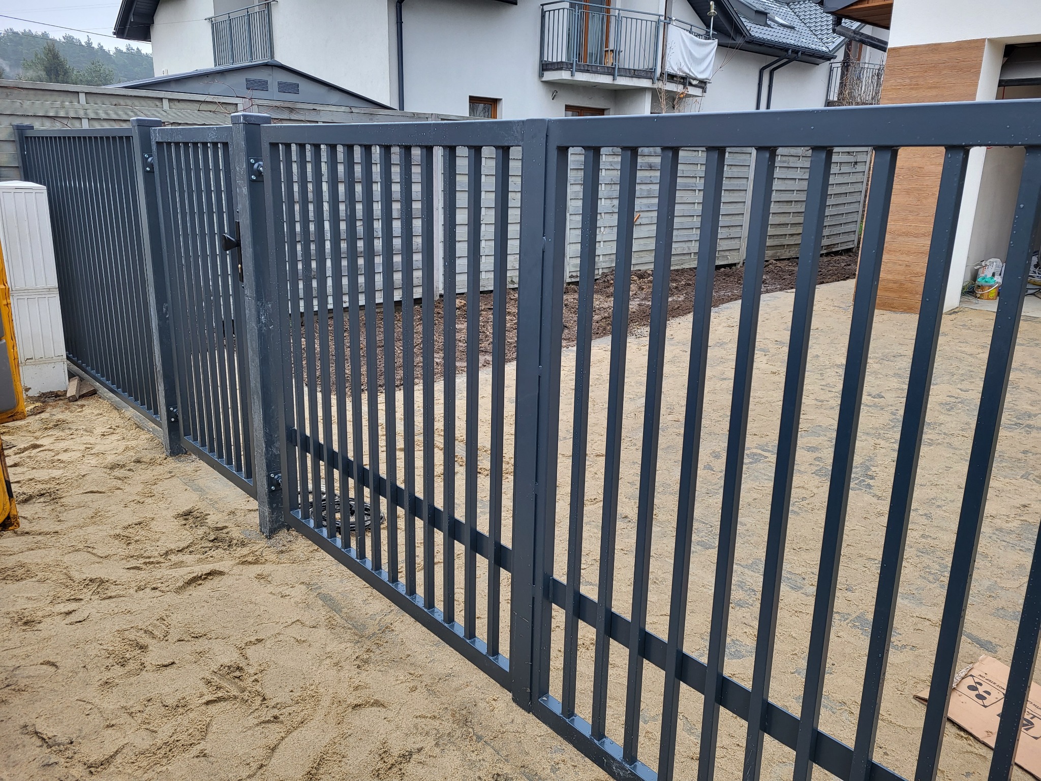 Nowoczesne, metalowe ogrodzenie w kolorze grafitowym z pionowymi prętami, świeżo zamontowane na piaszczystym podłożu, z widocznym budynkiem mieszkalnym w tle.