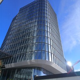 AMK Elektronic - Nowoczesny budynek biurowy z przeszkloną fasadą i widocznymi pracownikami w żółtych kamizelkach odblaskowych na niższych poziomach, sugerującymi prace instalacyjne lub wykończeniowe, z jasnym...