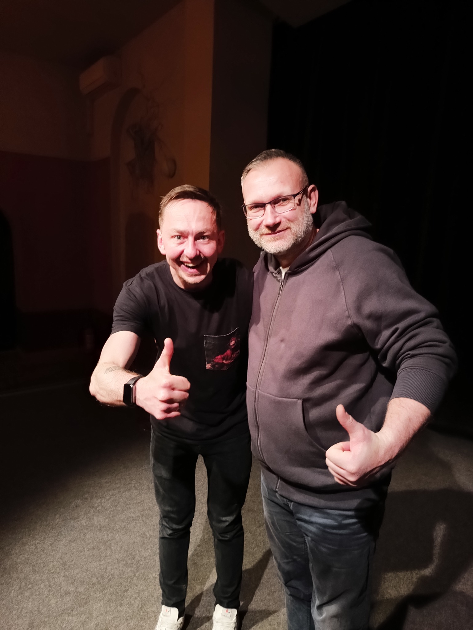 Dwóch uśmiechniętych mężczyzn, jeden w czarnej koszulce z kieszonką i tatuażem na przedramieniu, drugi w szarej bluzie z kapturem i okularach, obaj pokazują kciuki w górę na tle ciemnej ściany...